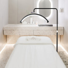 Load image into Gallery viewer, An adjustable LED lash light mounted over a spa bed, with a remote control visible in the frame.

